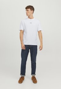 Hvid t-shirt med rund hals og korte ærmer, der har et lille brandlogo på brystet. Kombineret med mørkeblå denim jeans og brune sko.