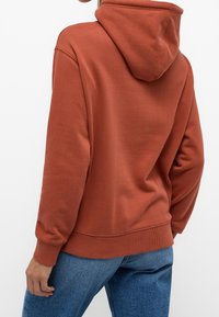 Sudadera de color óxido con una textura suave, bolsillo frontal tipo canguro, puños y dobladillo acanalados, y capucha con cordón ajustable. Combinada con jeans azules.