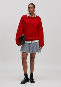 Roter übergroßer Pullover mit geripptem Saum, kombiniert mit einem weißen Hemd mit Kragen und schwarzen Streifen, dazu ein flauschiger grauer Rock und schwarze Slipper.