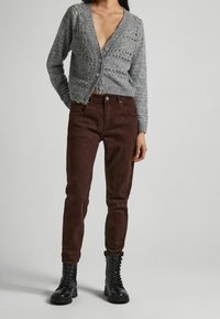 Cardigan grigio lavorato a maglia con trama aperta, jeans aderenti marrone scuro e stivali neri con lacci. Il cardigan presenta bottoni e scollo a V.