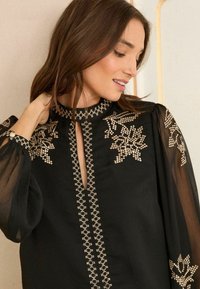 Melna blūze ar caurspīdīgām balonu piedurknēm, ar zeltītām izšuvumu ziedu rakstu un atslēgas formas kakla izgriezumu. Apstrādāta ar kontrastējošiem dūrieniem.