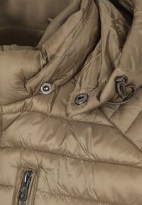 Beige pufferjack met een gladde, glanzende textuur. Voorzien van een ritsluiting, drukknoopsluiting en een quiltpatroon. Elastische manchetten en een capuchon. 