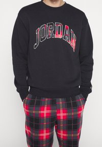 Person med en svart sweatshirt med texten "JORDAN" i röd rutig mönster och matchande röd, svart och grå rutiga byxor med händerna i fickorna.
