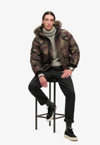 Pufferjakke i camouflage mønster med pelsfor i hætten, grå strikket turtleneck, sorte bukser og sorte sneakers med hvide såler.