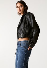 Giacca cropped in pelle nera con dettagli in zip e fibbia laterale, abbinata a jeans in denim blu a vita alta. Texture liscia, design aderente.