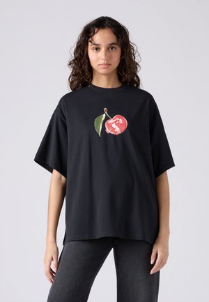 Mlada ženska z kodrastimi lasmi nosi ohlapno črno majico z rdečo češnjo in zelenim listom na prsih na enobarvni podlagi.