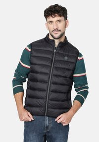 Chaleco acolchado negro con secciones acolchadas horizontales, cuello alto y cierre de cremallera. Lleva puesto un suéter de rayas verdes.