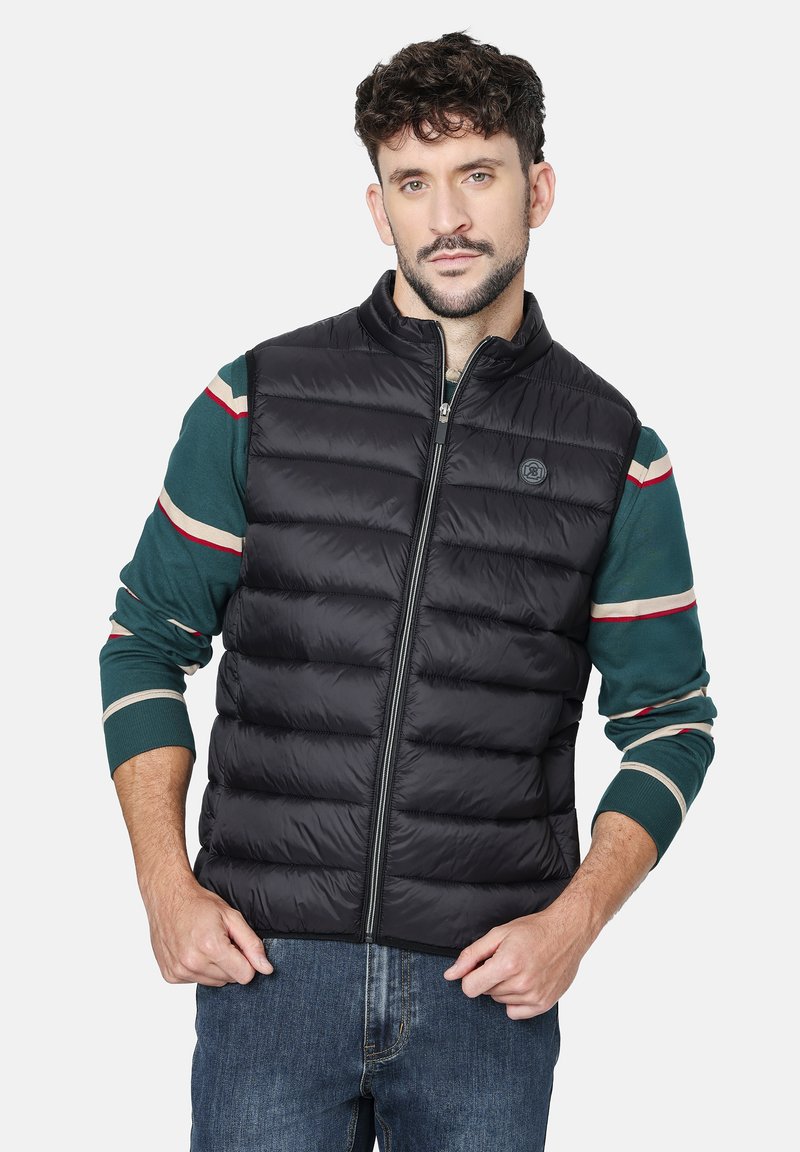 Chaleco acolchado negro con secciones acolchadas horizontales, cuello alto y cierre de cremallera. Lleva puesto un suéter de rayas verdes.