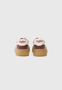 Des baskets rouges et crème présentent un design low-top, une semelle en caoutchouc robuste et des accents de marque au niveau du talon. Matériau lisse avec des panneaux contrastants.