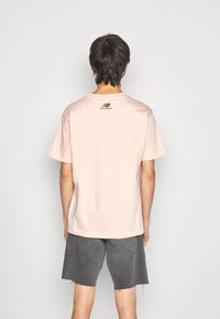 Camiseta de algodón de manga corta, de color rosa pálido, con cuello redondo y un detalle de logo negro en la parte superior de la espalda, combinada con pantalones cortos de mezclilla gris.