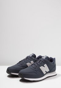 Zapatillas sintéticas de color azul marino con una suela blanca, que cuentan con un gran logo gris en forma de "N", diseño de cordones y acentos de malla texturizada alrededor del collar.