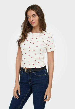 Woman with long brown hair wearing a white short-sleeve top with small red strawberries and dark blue jeans with a black belt.