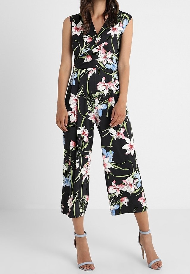 Femme portant une combinaison noire sans manches avec imprimé floral rose, blanc et bleu et jambes larges raccourcies, associée à des sandales grises à talons hauts.