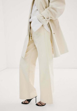 Personne portant un pantalon beige à jambes larges, un trench-coat assorti, une chemise blanche et des sandales noires à talons hauts, debout sur un sol blanc.