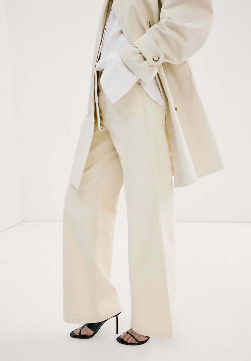 Personne portant un pantalon beige à jambes larges, un trench-coat assorti, une chemise blanche et des sandales noires à talons hauts, debout sur un sol blanc.