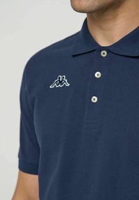 Férfi tengerészkék galléros pólóban, három gombbal és a bal mellrészen fehér hímzett logóval, amely két háttal egymásnak ülő alakot ábrázol.