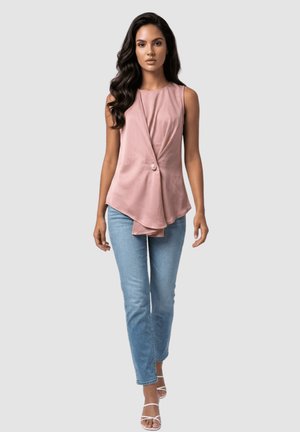 Mujer con cabello largo y oscuro que lleva una blusa asimétrica sin mangas de color rosa, vaqueros azul claro y sandalias blancas con tiras sobre un fondo liso.