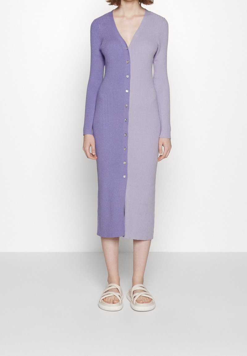 Femme portant une robe midi côtelée, boutonnée, à manches longues, divisée verticalement en deux nuances de violet avec des sandales blanches.