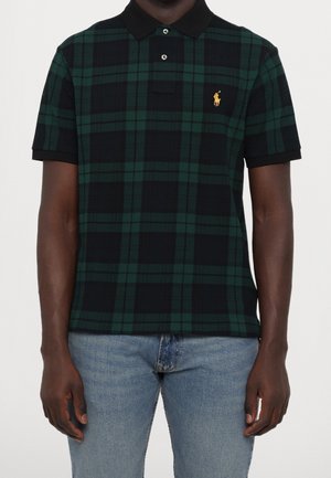 Man draagt een groen-zwart geruit poloshirt met korte mouwen en een zwarte kraag, gecombineerd met lichtblauwe spijkerbroek, staand tegen een witte achtergrond.