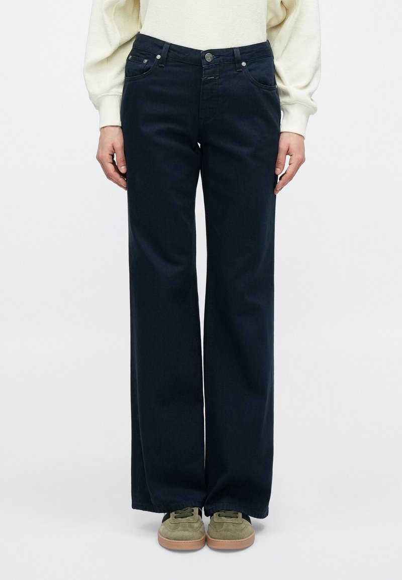 Tumši zili plašām kājām džinsi no kokvilnas ar priekšējām kabatām un pogu aizdari. Saskaņoti ar zaļām zamšāda kurpēm kontrastam.