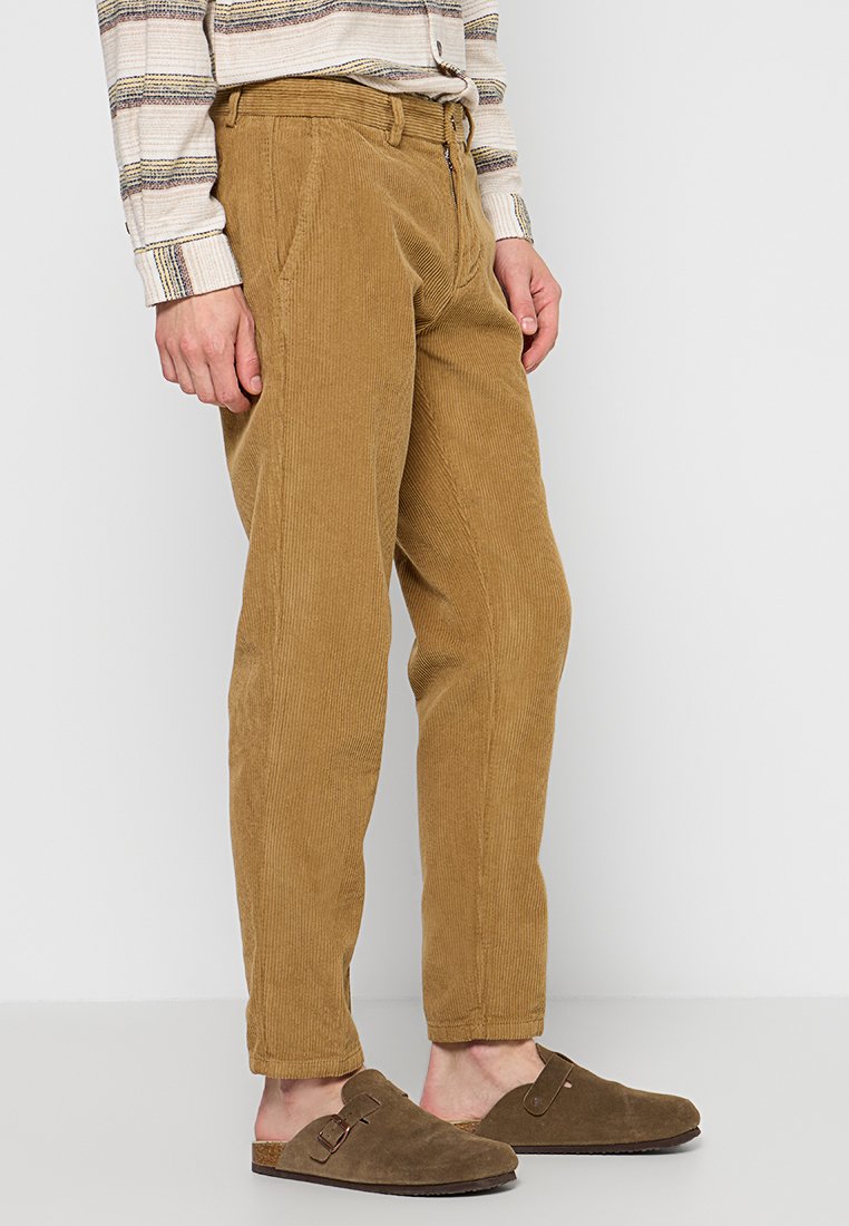 Pantaloni in corduroy di un caldo colore marrone con coste verticali visibili, dotati di tasche laterali e una vestibilità comoda, abbinati a scarpe slip-on marroni.