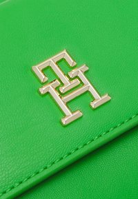 Zelená kožená kabelka s zlatým geometrickým logem. Hladký povrch s precizním švem po okrajích. Kompaktní, moderní design.