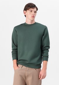 Sweatshirt verde escuro com decote redondo, mangas longas e punhos canelados. Textura suave, combinado com calças claras em risca de giz.