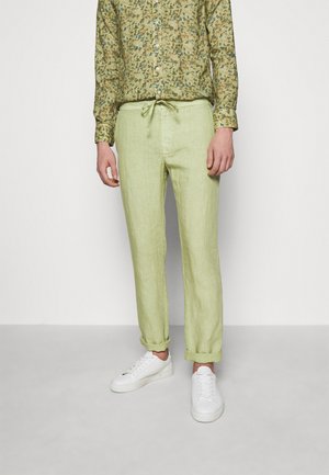 Homme portant un pantalon vert clair à cordon, ourlets roulés, baskets blanches et une chemise verte à motifs à manches longues, debout devant un fond uni.