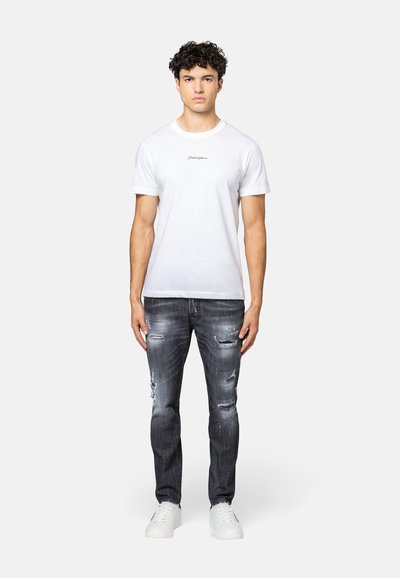 Witte katoenen t-shirt met geborduurde zwarte tekst, gecombineerd met grijze distressed jeans en witte sneakers, gedragen door een staande mannelijke model.
