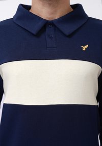 Man wearing navy blue polo shirt with a wide white horizontal stripe and a small yellow bird logo on the left chest.