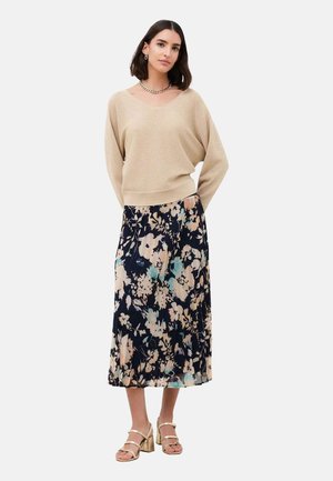 Femme portant un pull beige, une jupe midi fleurie et des sandales à talons beiges, debout les mains derrière le dos sur un fond uni.