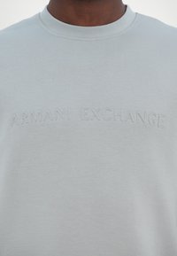 Light gray crewneck sweatshirt featuring embossed text "ARMANI EXCHANGE" on the front. Fabric appears soft with a smooth texture.