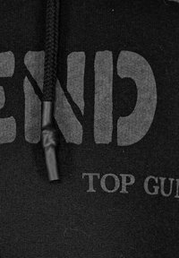 Svart hoodie med stor, fet, grå text. Har en texturiserad, flätad dragsko och en slät tygfinish. Utsmyckad med tryckta grafiker.