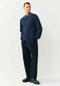 Marineblaues Langarm-Polo mit kontrastierendem Kragen, kombiniert mit lockeren schwarzen Hosen und schwarzen Schuhen. Glatte Stofftextur, entspannte Passform.