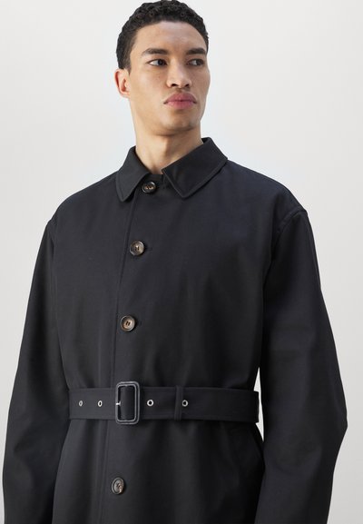 Černý kabát s páskem v pase, který má límec, velké knoflíky a hladkou texturu, navržený v rovné siluetě pro minimalistický vzhled.