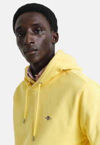Gele katoenen hoodie met trekkoord en logo op de borst, gelaagd over een roze en witte gestreepte shirt. Glad textuur, casual pasvorm.