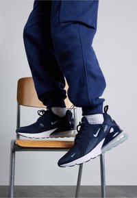 Marinträningsskor med ovandel i mesh och syntetmaterial, vita accenter, luftkuddesula, i kombination med marinblå byxor, placerade på en stol.