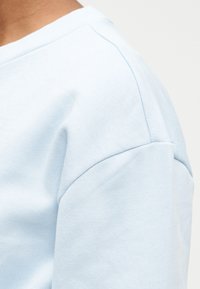 Ανοιχτό μπλε φούτερ με ομαλή υφή, στρογγυλή λαιμόκοψη και μανίκια ραγκλάν με λεπτομέρειες ραφής.