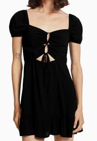Musta mekko lyhyillä puffi-hihoilla, neliönmuotoisella kaula-aukolla ja etu-leikkauksella. Sisältää koottua yksityiskohtaa ja leveän helman, jossa on röyhelöreuna.