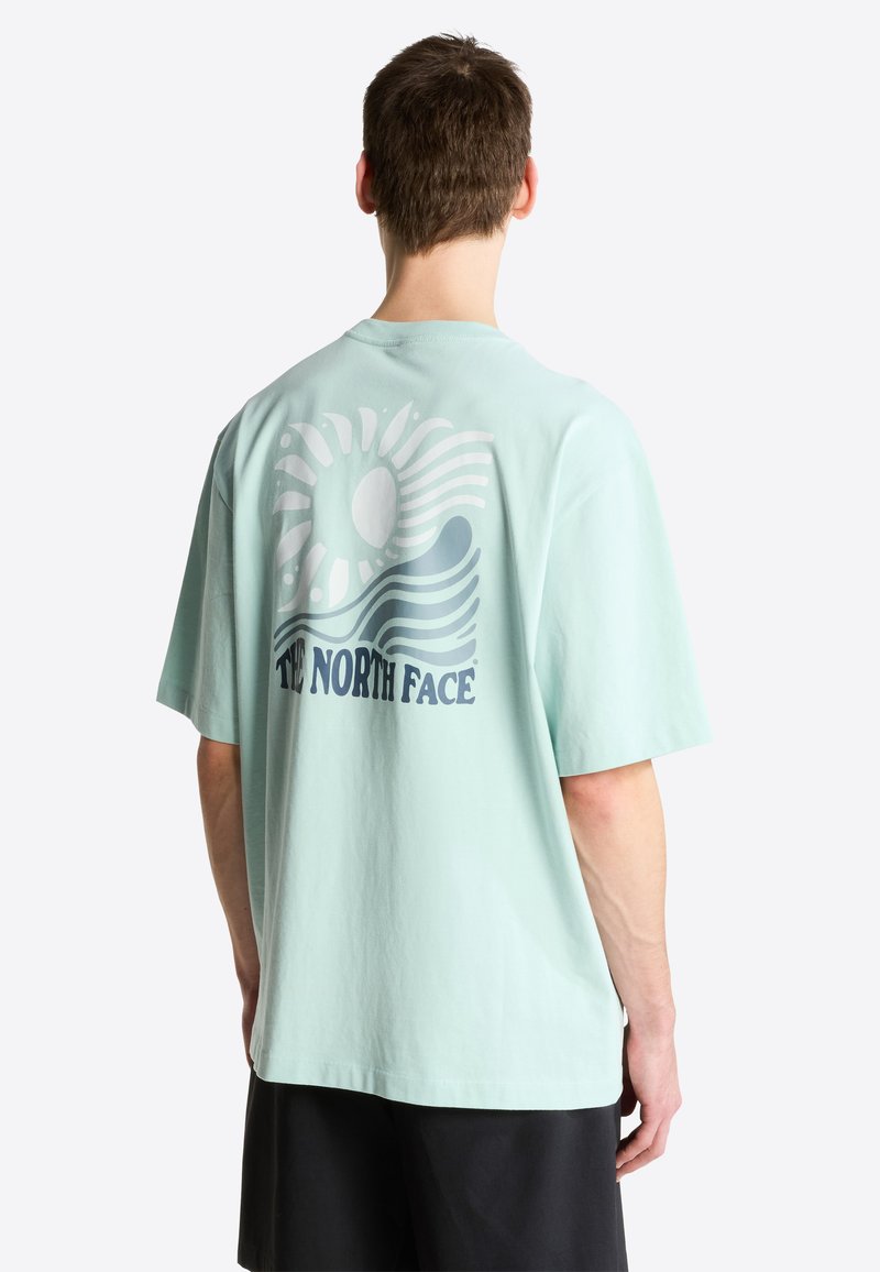 Ein Mann, von hinten gesehen, trägt ein hellblaues T-Shirt mit Sonnen- und Wellengrafik sowie dem Text "THE NORTH FACE" auf dem Rücken, kombiniert mit schwarzen Shorts.