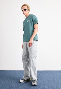 Zöld pamut póló "DICKIES" logóval, szürke cargo nadrág oldalzsebekkel, és szürke sportcipő fekete és sárga részletekkel.