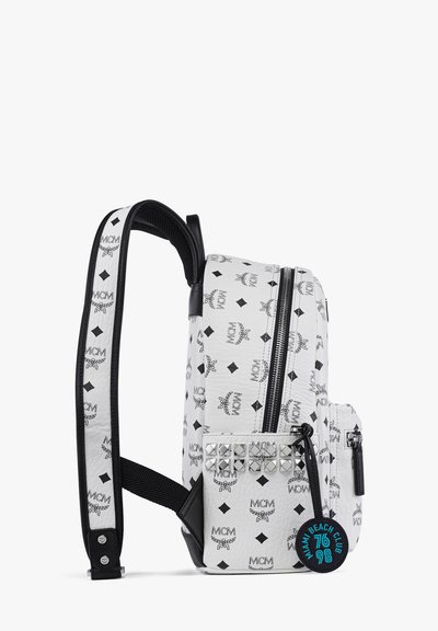 Weißer Rucksack mit schwarzen MCM-Logos, Reißverschluss-Hauptfach, Vordertasche mit silbernen Nieten, verstellbare schwarze Träger, runder blauer Anhänger.