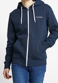 Sudadera con cremallera en color navy con cordones blancos, logo de Calvin Klein en el pecho, hecha de una tela suave, con un bolsillo canguro delantero y puños de canalé.