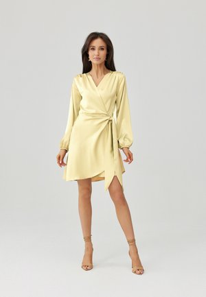 Kobieta w jasnożółtej satynowej sukience typu wrap z długimi rękawami i wiązaniem z boku, w połączeniu z beżowymi sandałami na wysokim obcasie, stojąca na jednolitym tle.