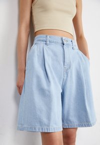 Shorts en denim bleu clair avec une coupe décontractée, des plis à l'avant et une fermeture à bouton. Dispose de poches latérales et d'un bord ourlé.
