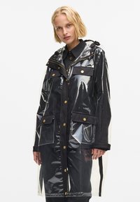 Impermeable transparente con acentos de tela negra, con capucha, herrajes dorados y múltiples bolsillos delanteros. Corte largo y recto.