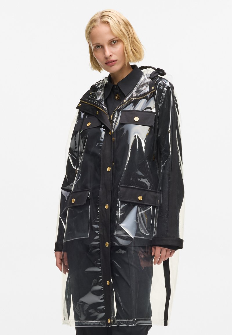 Impermeable transparente con acentos de tela negra, con capucha, herrajes dorados y múltiples bolsillos delanteros. Corte largo y recto.