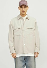 Beige corduroy shirt jacket with two chest pockets, button closure, and a relaxed fit, worn over a white shirt and blue jeans.