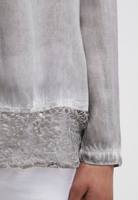 Blouse grise en tissu transparent avec bordure en dentelle à l'ourlet. La manche est retroussée avec un détail de bouton, mettant en valeur la texture et le design.