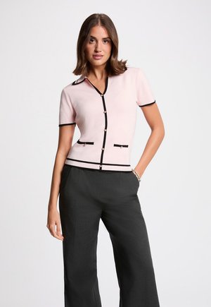 Kvinde står med den ene hånd i lommen iført lyserød kortærmet cardigan med sort kant og mørke bukser med brede ben.